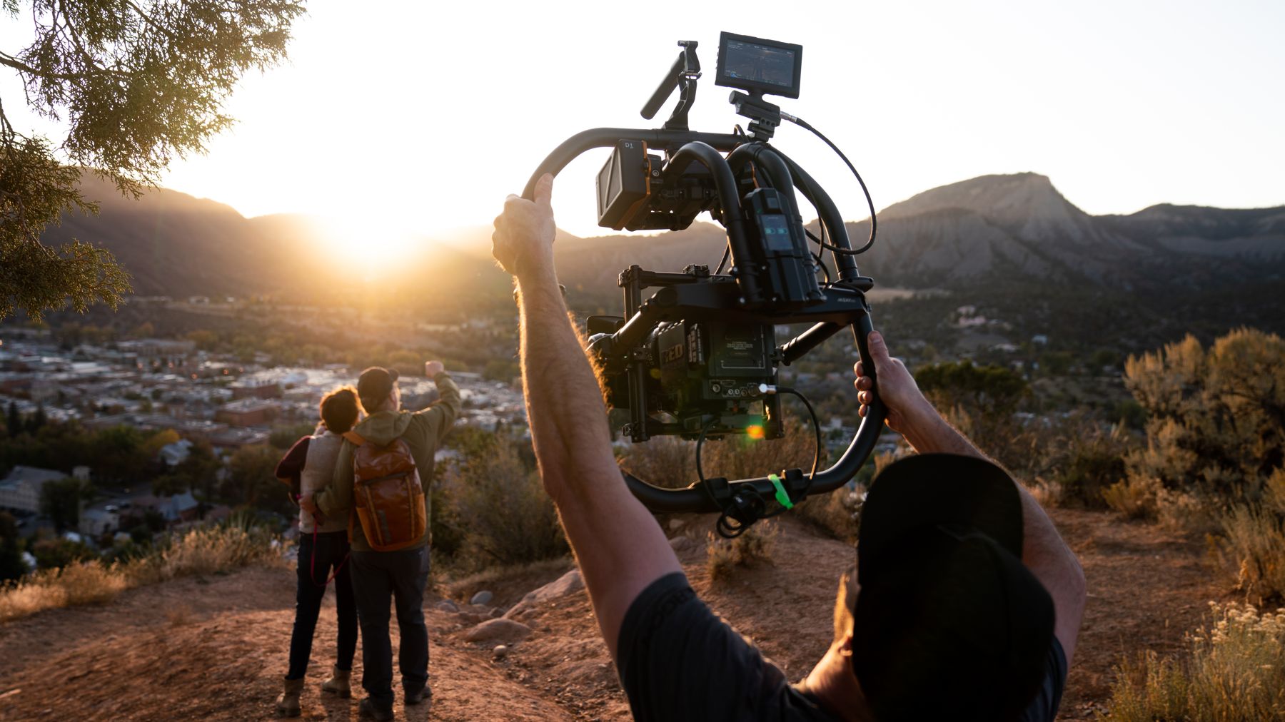 A camera operator films a couple overlooking a mountain town at sunset.